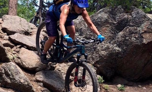 A mountain biker navigates rocky terrain among large boulders in a forest.