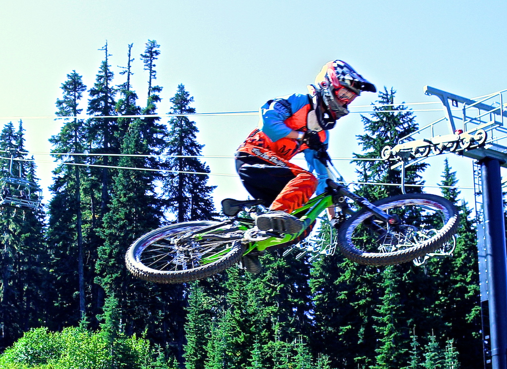 A mountain biker airborne off a jump with coniferous forest and a chairlift in the background.