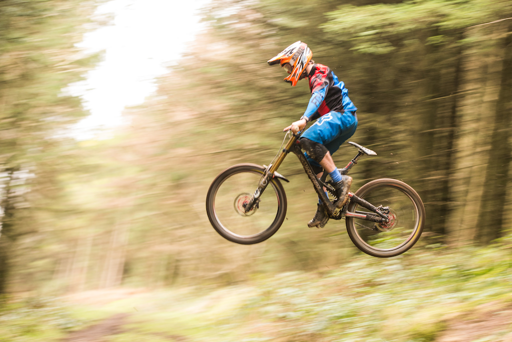 A mountain biker launching airborne over a gap in a wooded trail.