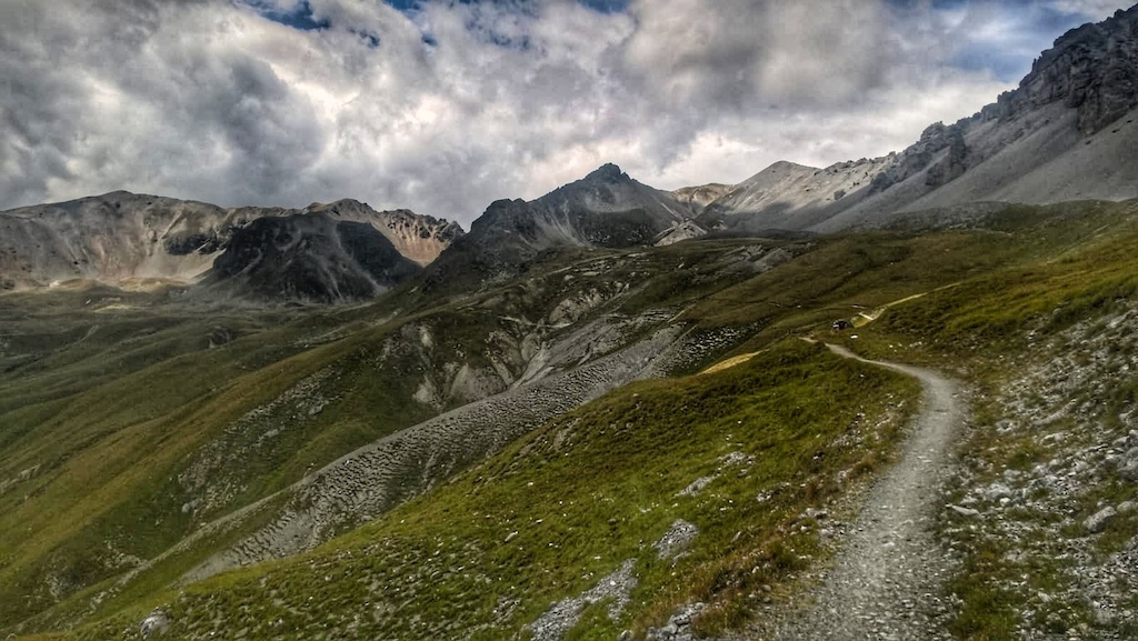 Winding gravel trail descending through alpine meadows with jagged mountain peaks and colorful slopes.