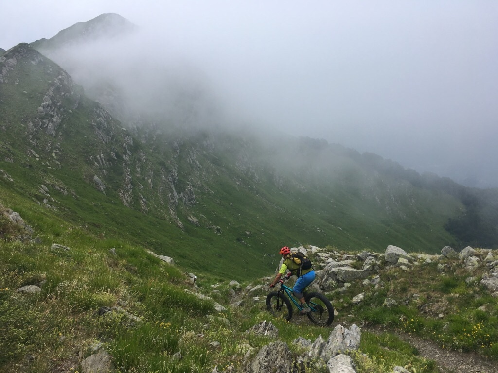 A mountain biker riding rocky alpine terrain with green slopes and misty peaks above.
