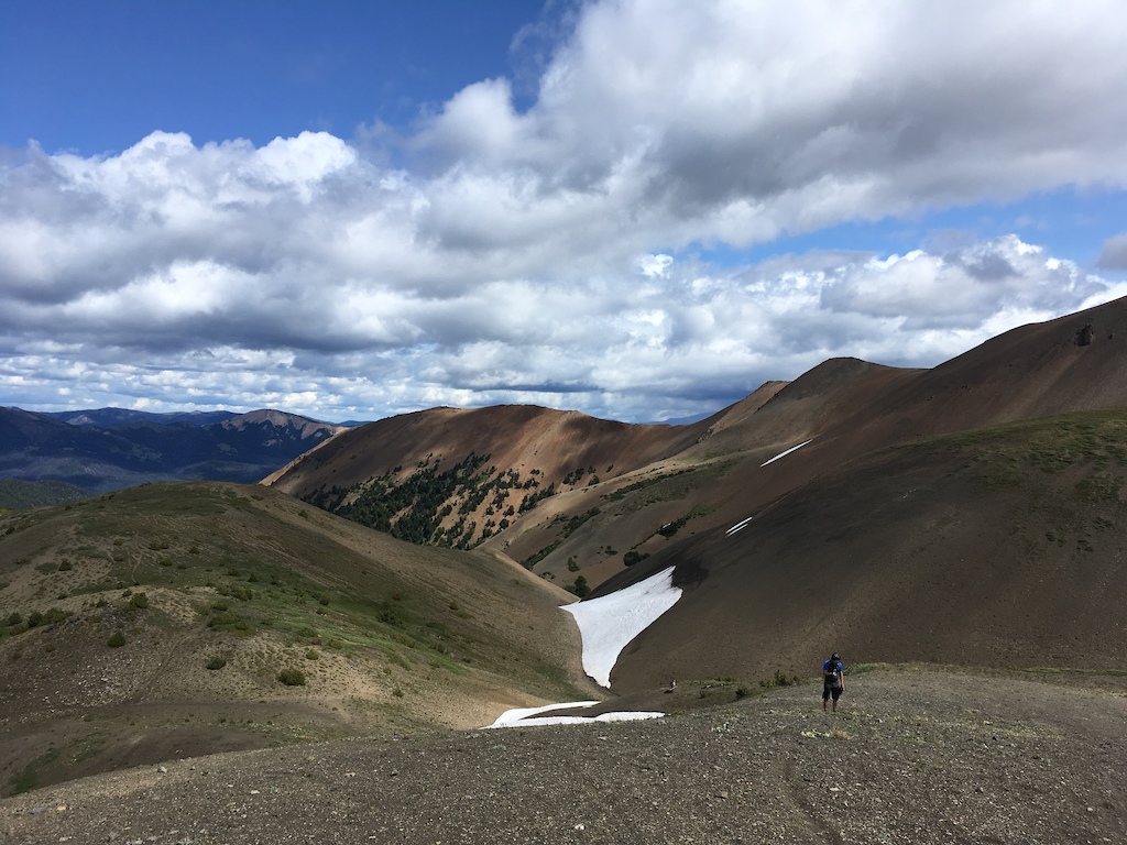 A hiker on alpine terrain with patches of snow and distant mountain views.