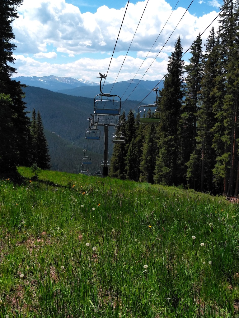 A ski lift overlooks a forested valley with distant snow-capped mountains under blue sky.