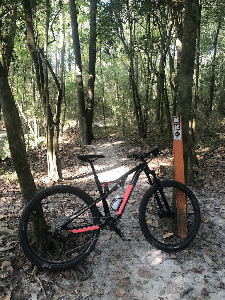 A mountain bike with red accents parked on a forest trail next to a brown trail marker sign.