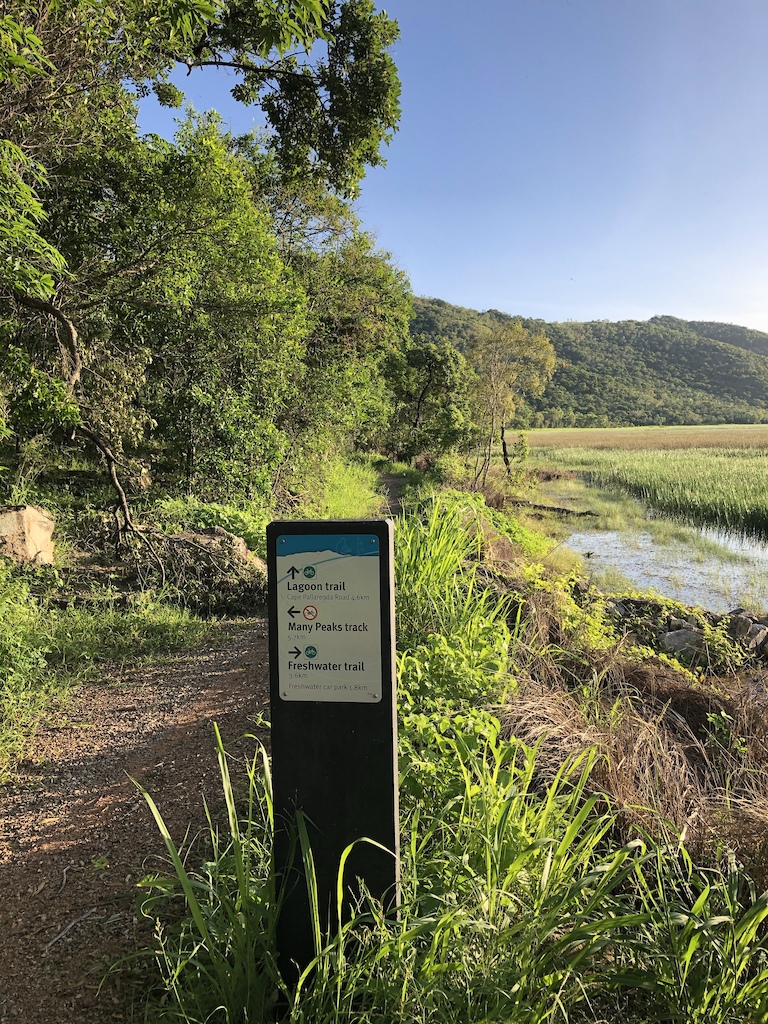 A trail sign at a fork with a lagoon and hillside visible in background.