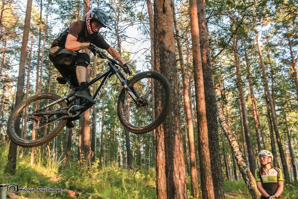 A mountain biker jumps over a root feature in a pine forest while a spectator watches.