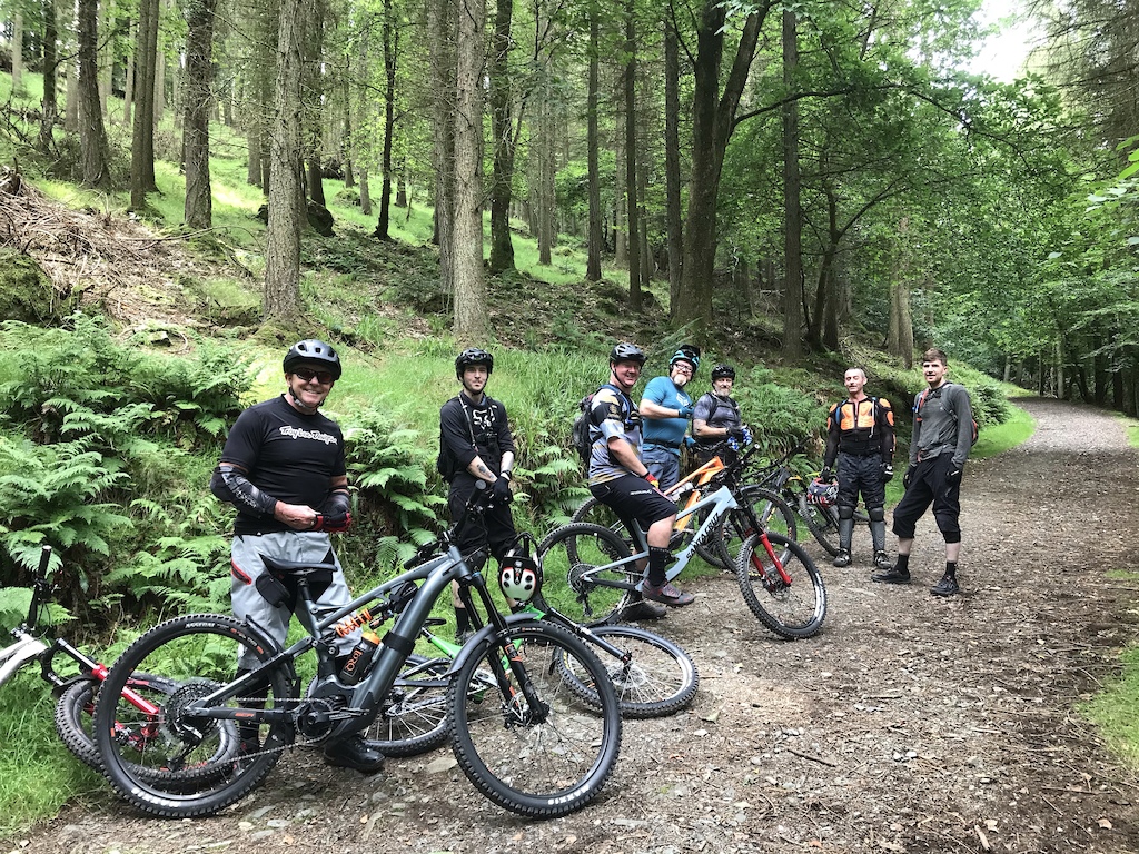 A group of mountain bikers gathered on a dirt trail in a forest, posing for a photo.