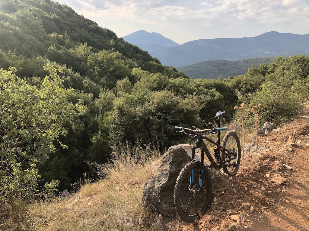 A mountain biker with a gravel bike paused on a rocky trail overlooking forested hills and distant mountains.