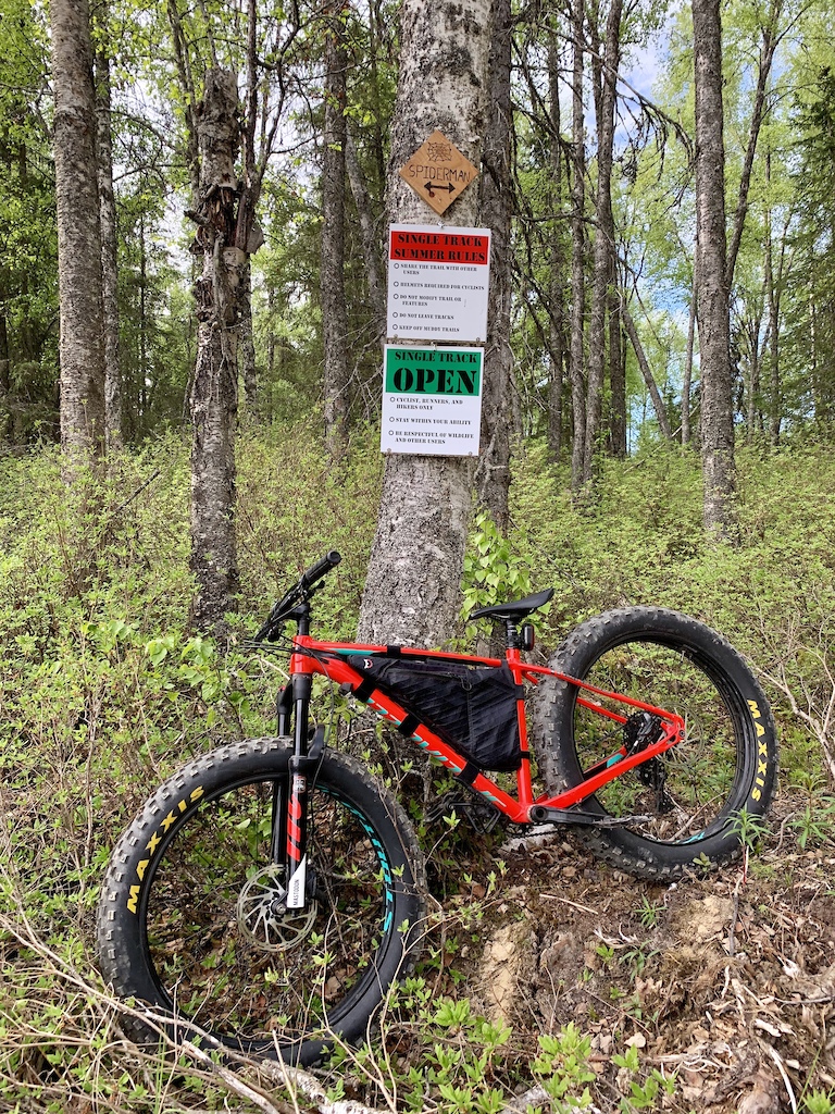A red mountain bike leaned against a tree with trail signs in a leafy forest.