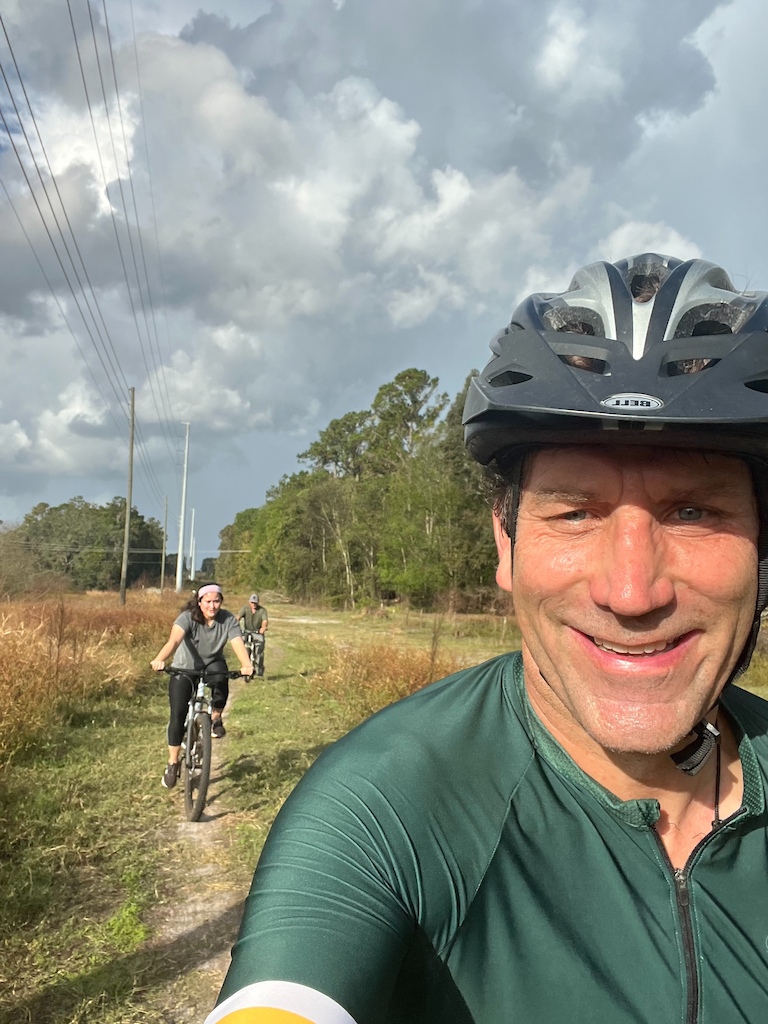 A cyclist in a selfie with another rider visible behind on a trail through open scrubland.