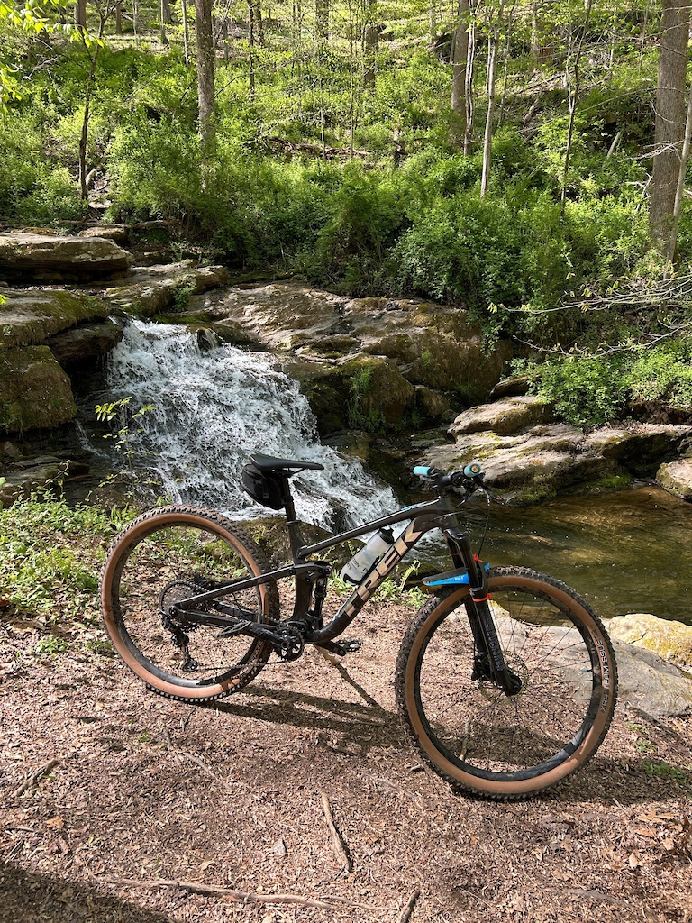 A gravel bike rests on dirt beside a small waterfall in a rocky creek flowing through forest.
