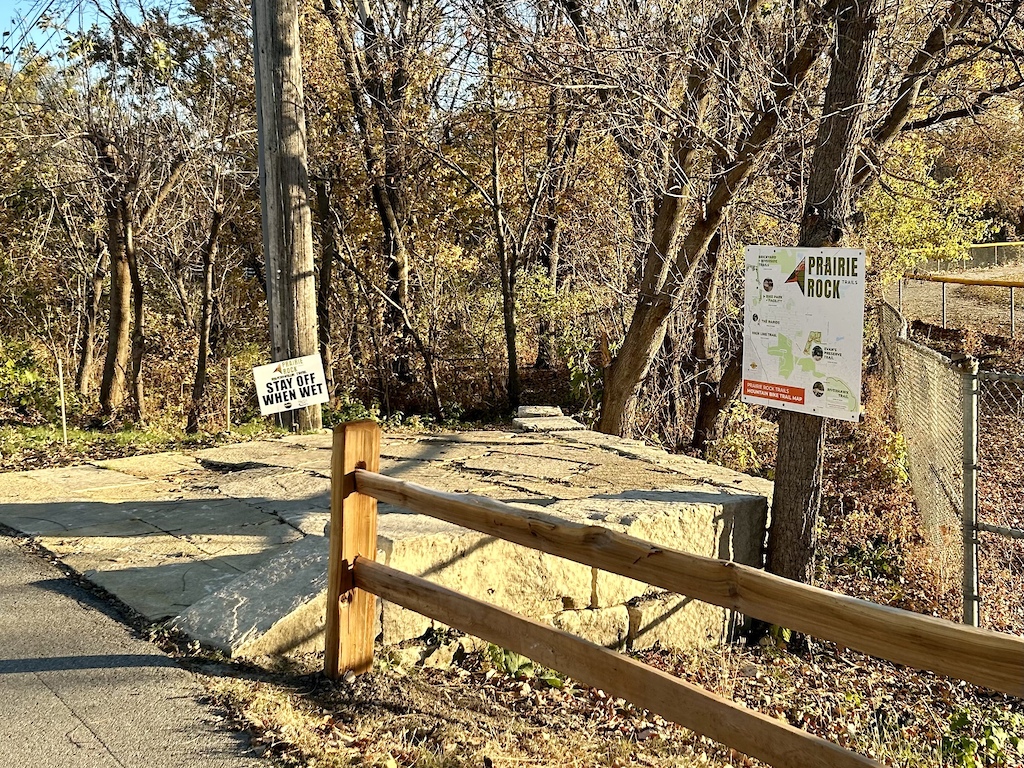 A trail sign and wooden fence at a forest trailhead entrance with fall foliage.