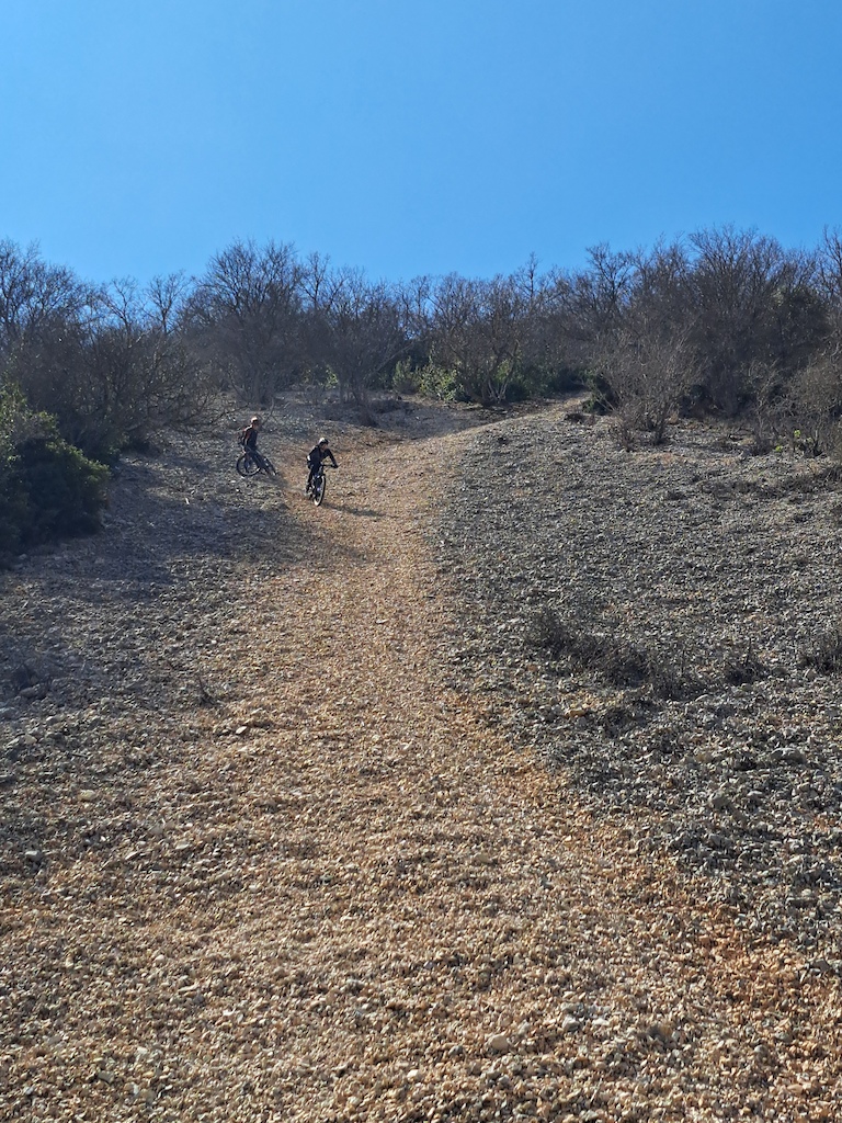 Two mountain bikers riding on a rocky, sparsely vegetated gravel slope under clear blue sky.