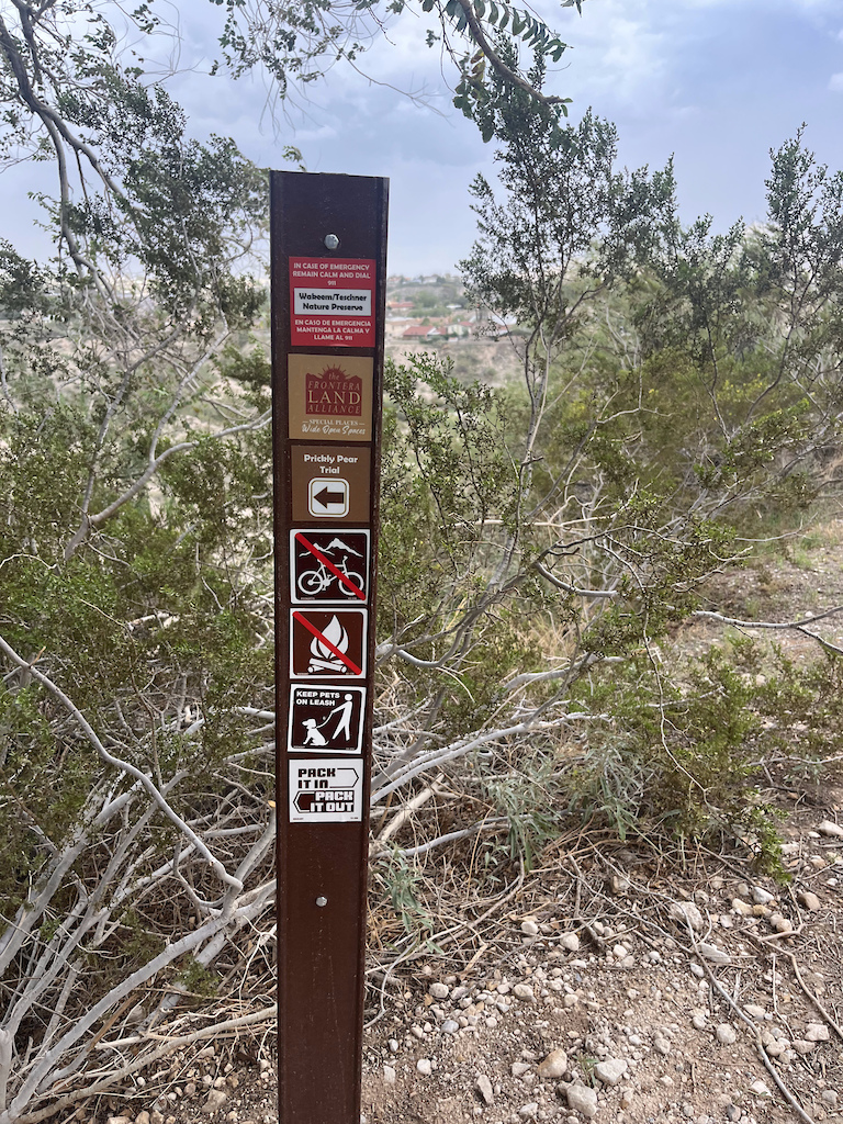 A brown trail sign post with hiking and fire icons in arid shrubland overlooking a distant valley.