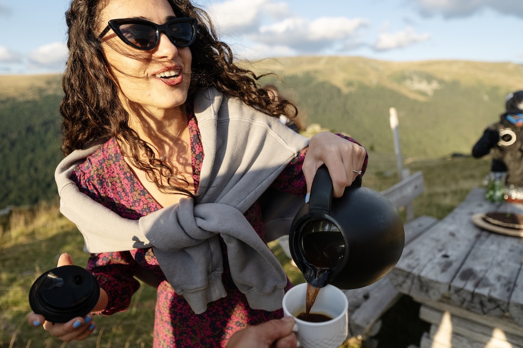 A woman posing with a coffee pot and cup, off-trail near a mountain hut.