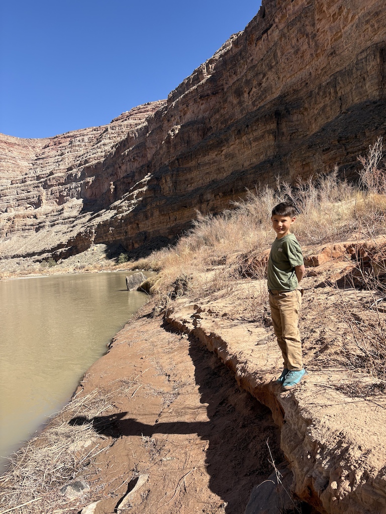 A hiker stands by a muddy river at the base of tall canyon walls with layered rock formations.