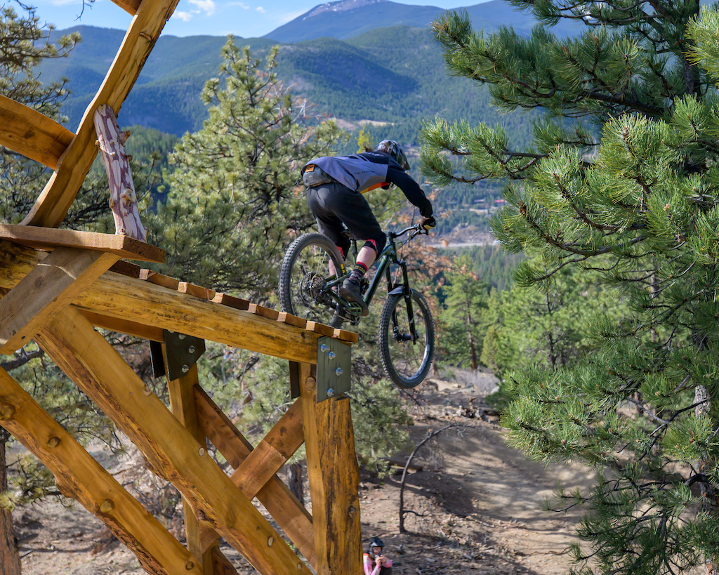 A mountain biker riding across a wooden feature with mountain valley views and pine forest below.
