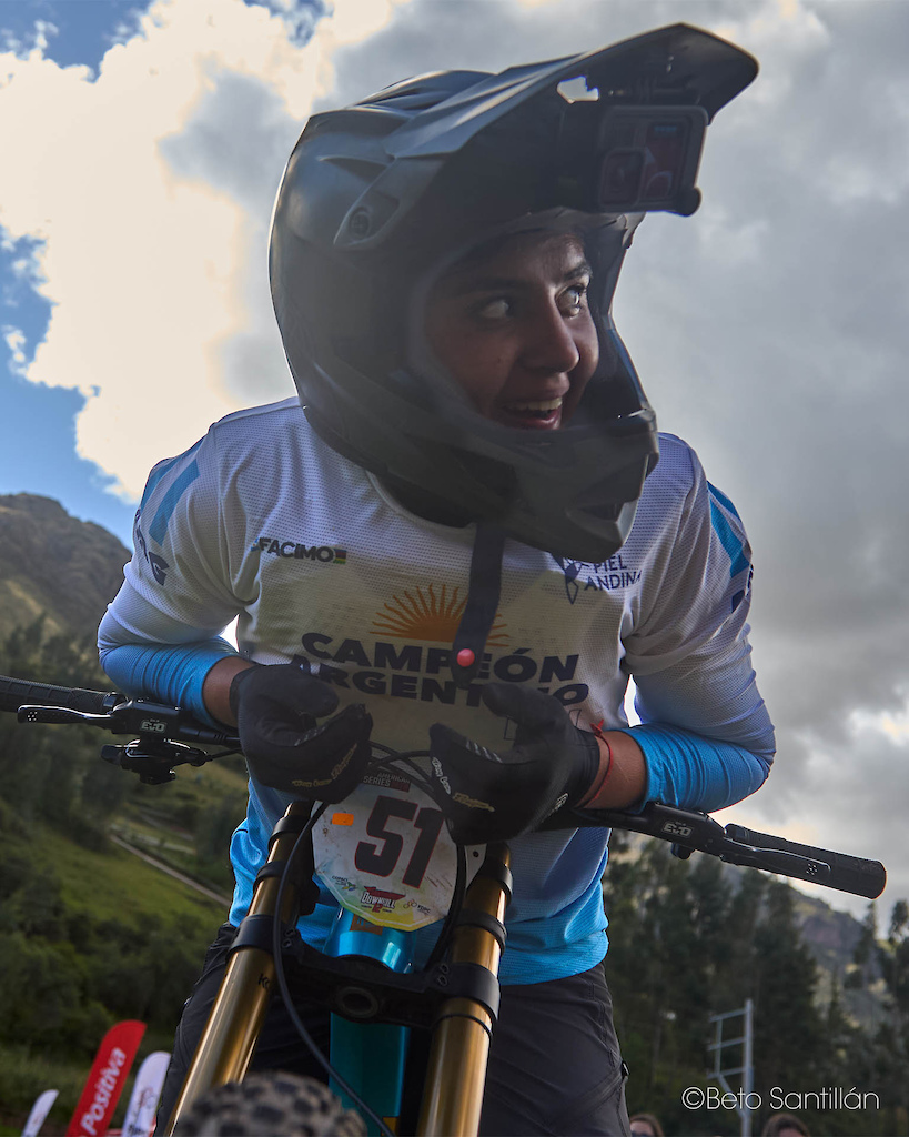Catalina Calderón — This is the face of a champion in the exact moment she realizes she has just conquered Santísimo DH 12. Helmet still on, breath still racing, eyes searching for the scoreboard that will confirm what her heart already knows. The sky breaks open above the Sacred Valley, the Andes stand witness, and number 51 — Campeona Argentina — sits at the bottom of the mountain having left absolutely everything on that track. This expression is worth more than a thousand words. This is what winning feels like from the inside