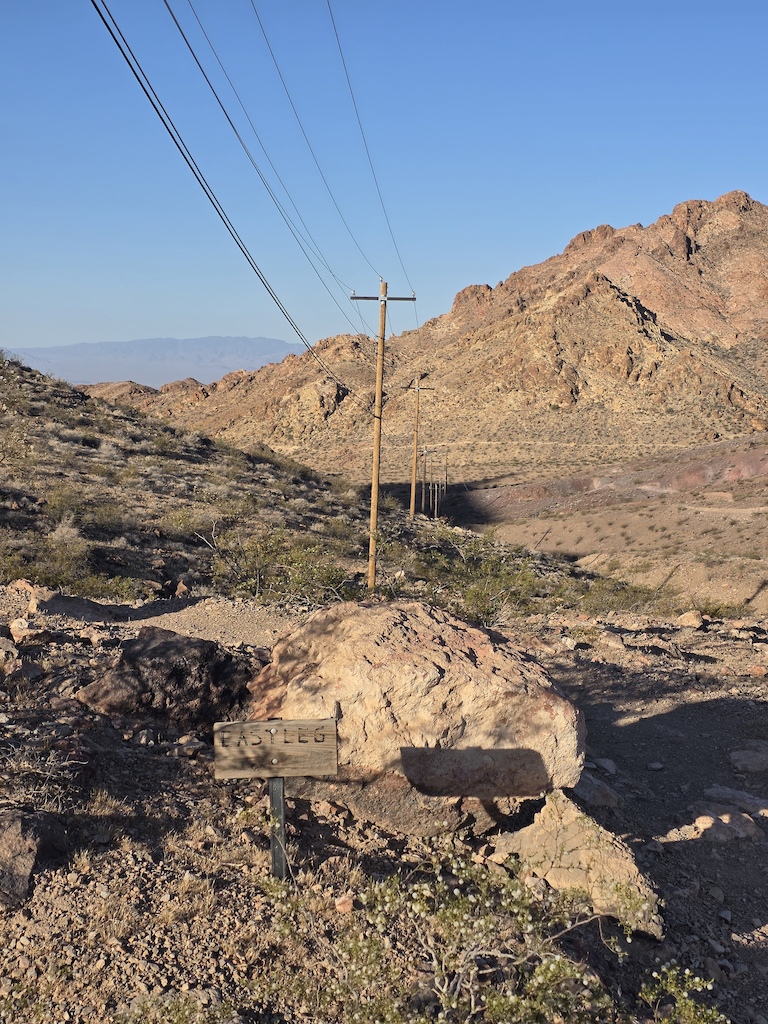 A trail marker sign on rocky desert terrain with power lines and mountains.