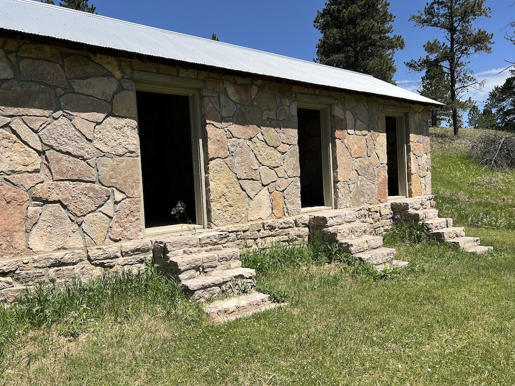 A historic stone building with multiple doorways and a metal roof surrounded by grass and trees.