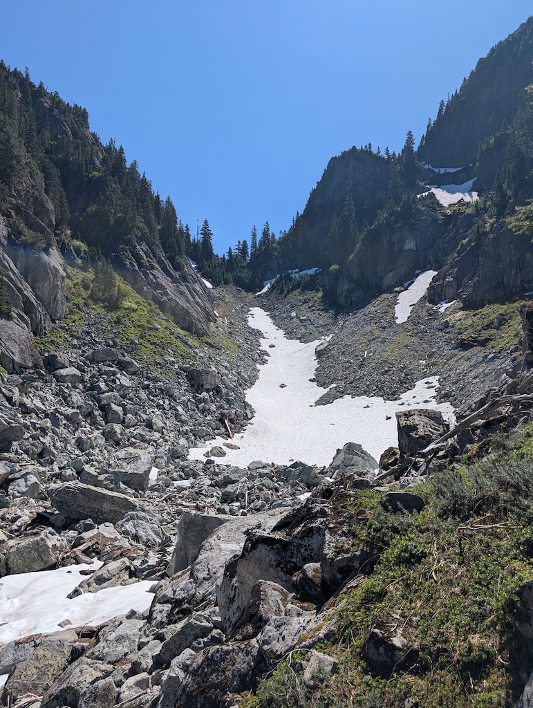 A canyon with rocky slopes, patches of snow, and evergreen trees under a clear blue sky.