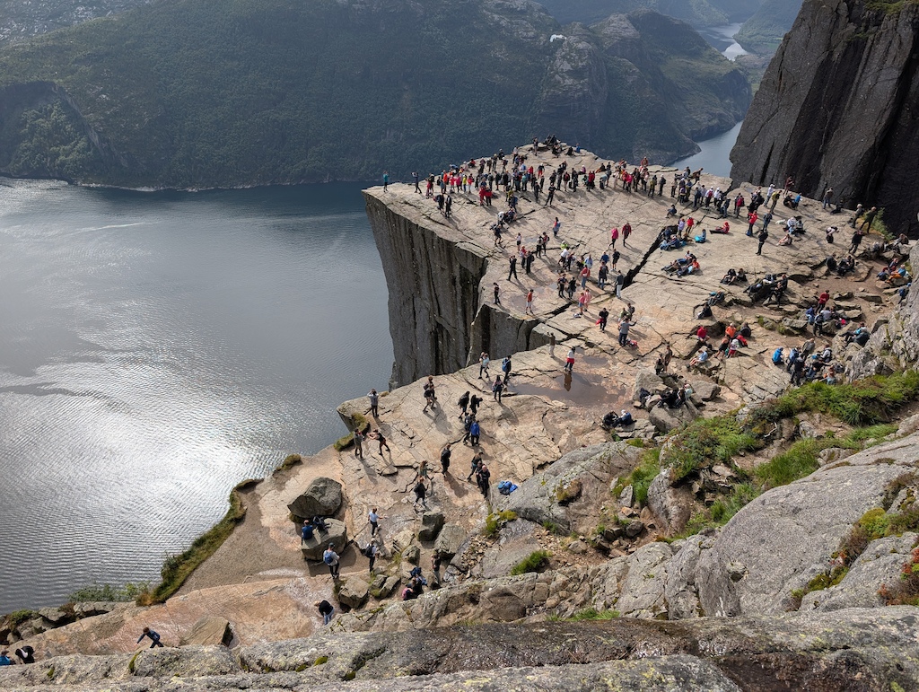 People hiking on a rocky plateau overlooking a fjord with mountains and water below.