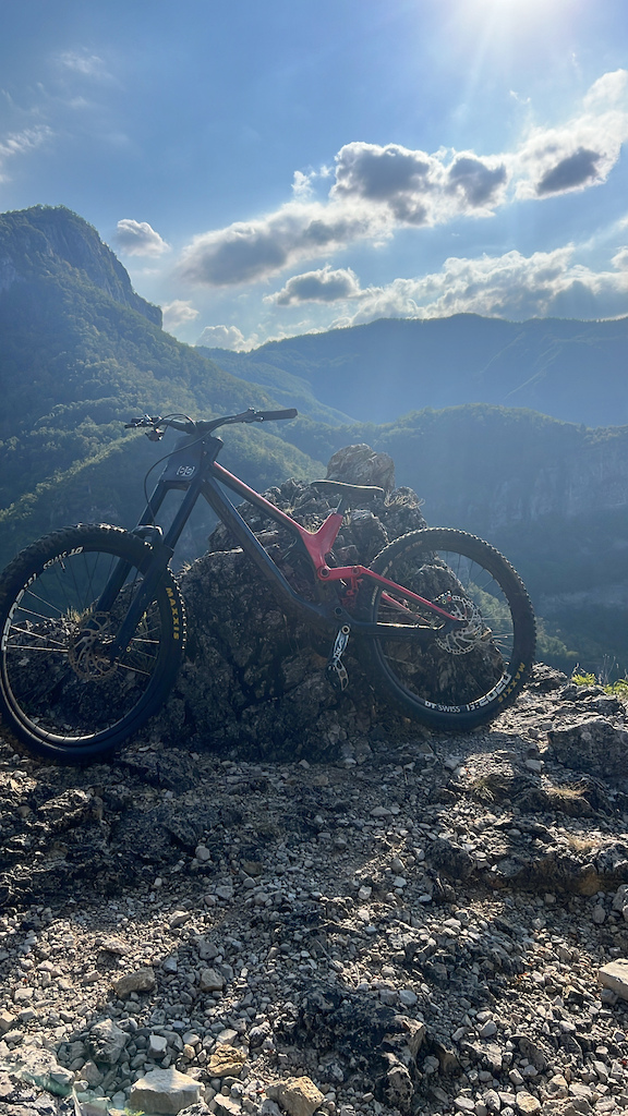 A red and black mountain bike resting on rocky terrain with mountain valley views and clouds.