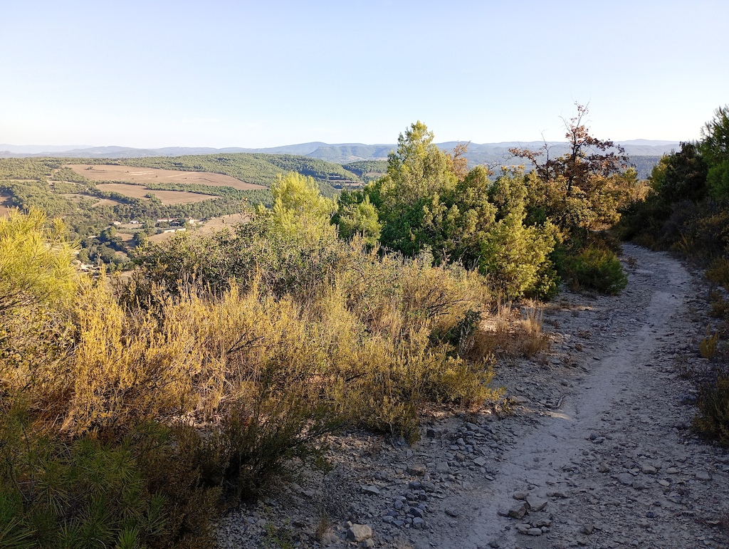 A rocky trail descending through desert scrub with distant mountains and valleys under clear sky.