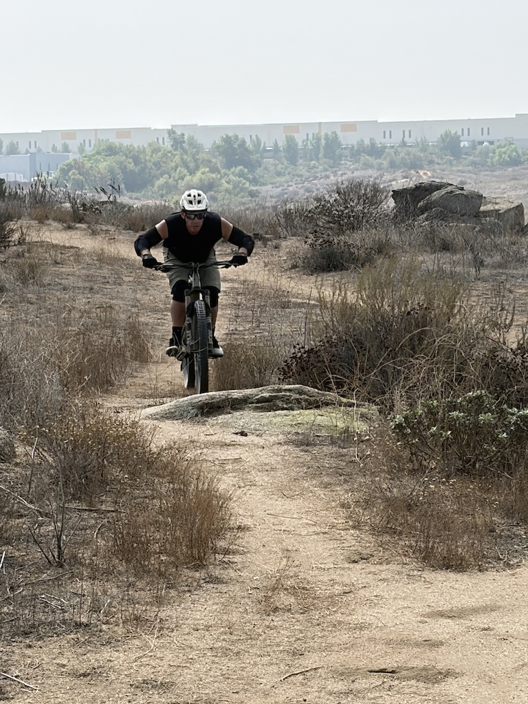 A mountain biker riding on a dirt trail through scrubland with buildings visible in the distance.