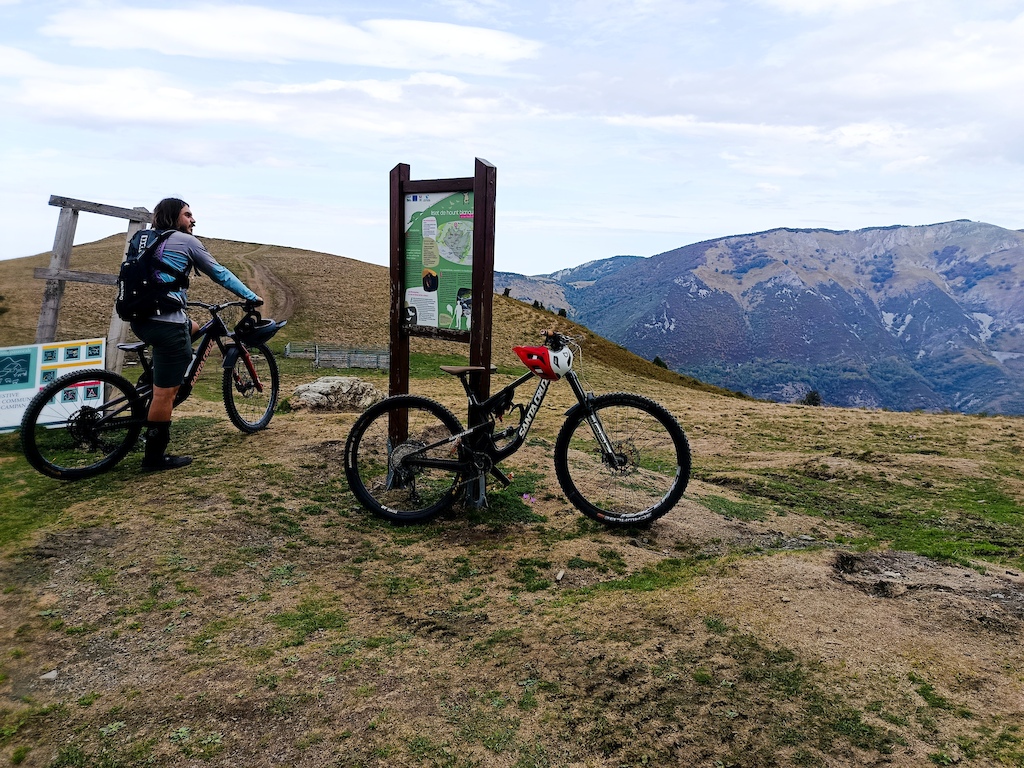 Two cyclists standing at trailhead with map sign and mountain views.