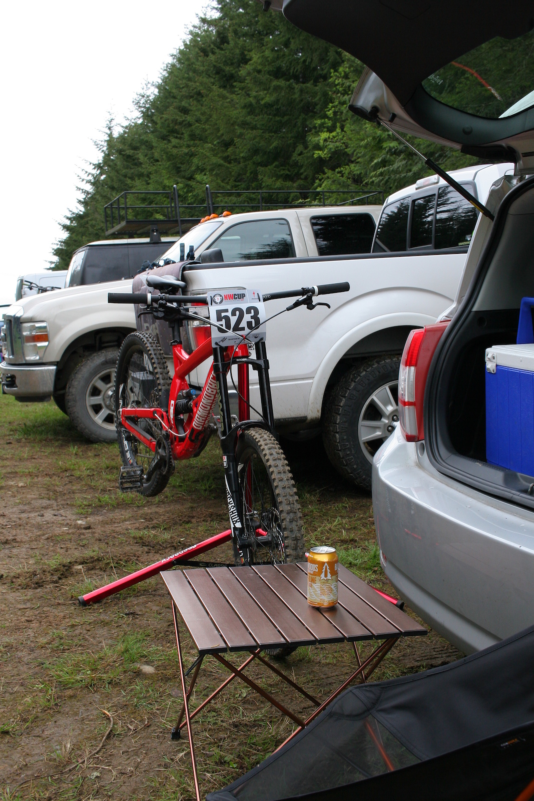 A red mountain bike with race number 523 parked at a parking area with vehicles and trees.