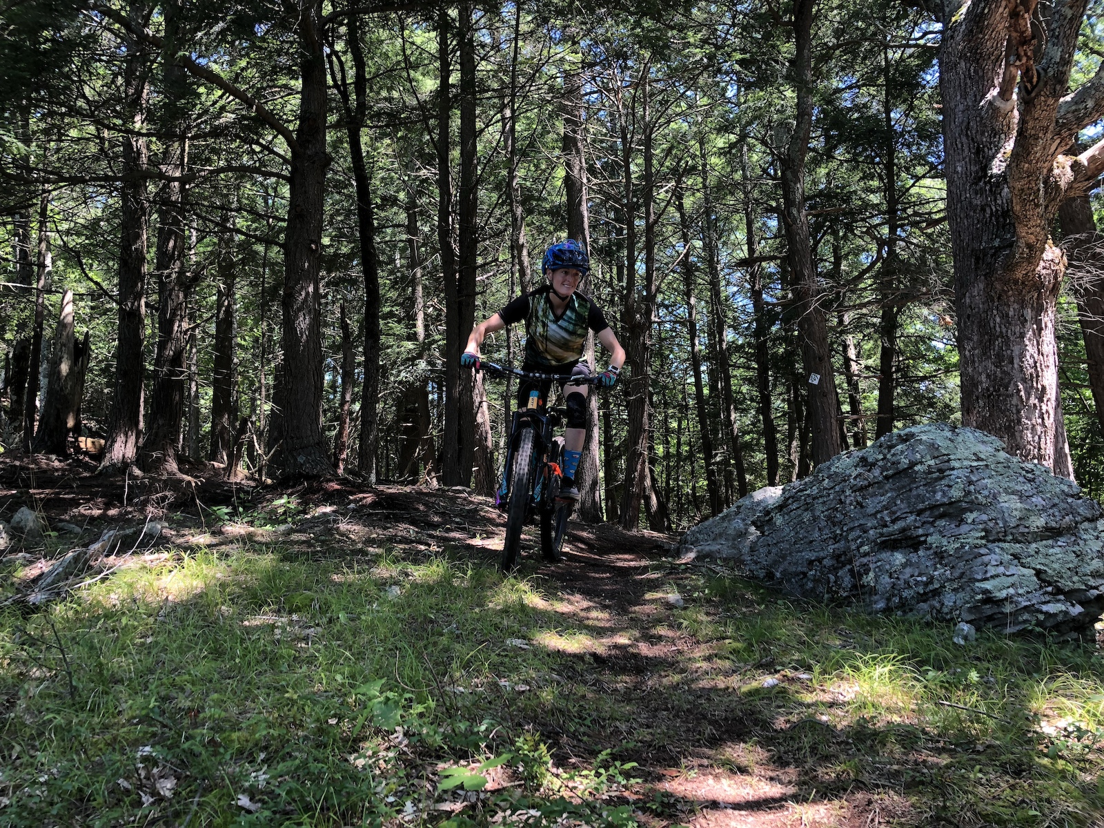 A mountain biker on a rocky forest trail with trees and boulders.
