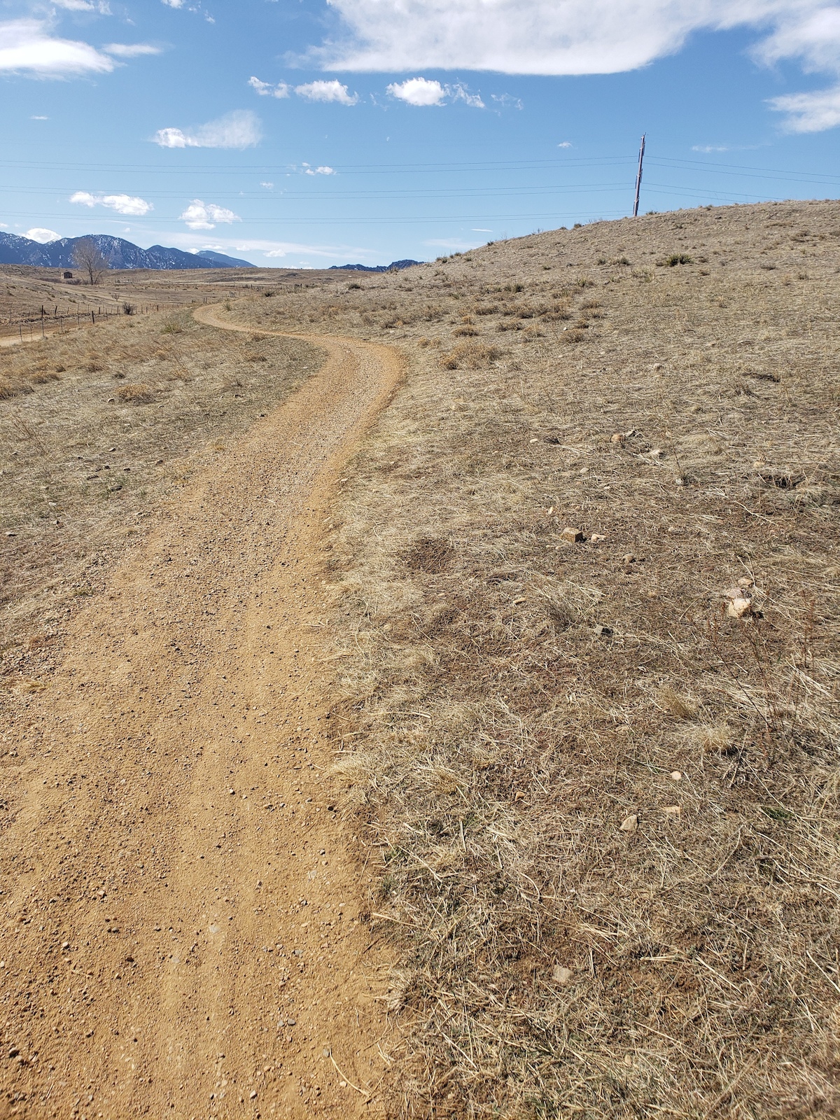 A dirt trail winds through sparse desert scrubland with distant mountains under blue sky.