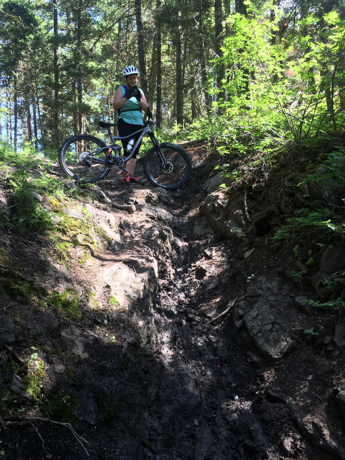 A mountain biker with a bike on a rocky trail with large boulders in a green forest.