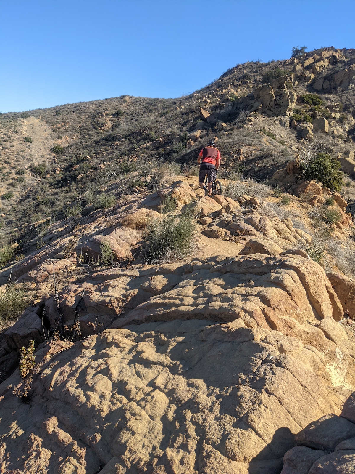 A mountain biker riding over rocky slabs on a desert trail with rock formations visible.