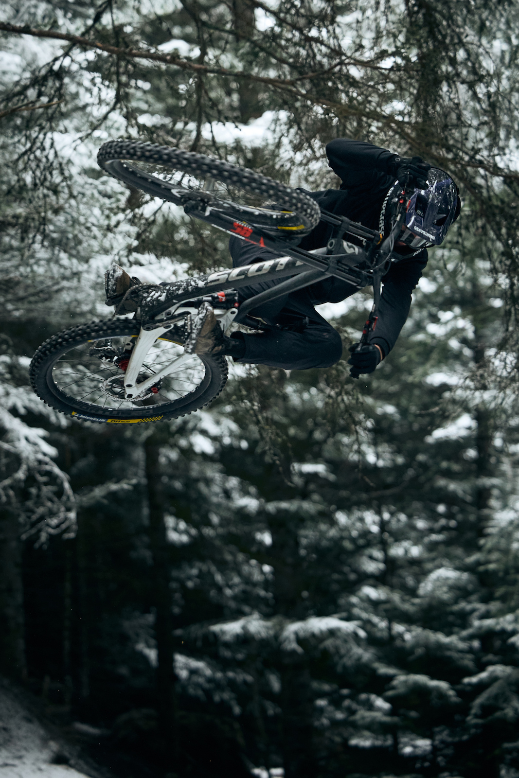 A mountain biker airborne mid-trick in a snowy forest with evergreen trees.