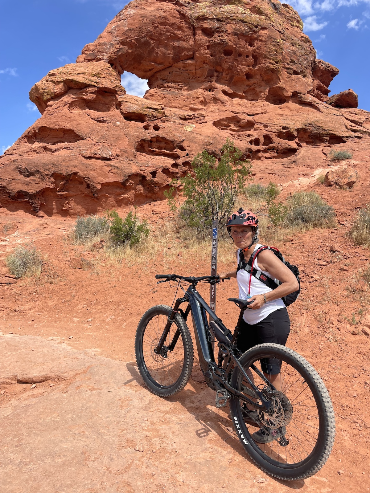 A female mountain biker standing with her bike on red desert terrain beneath a distinctive rock arch formation.