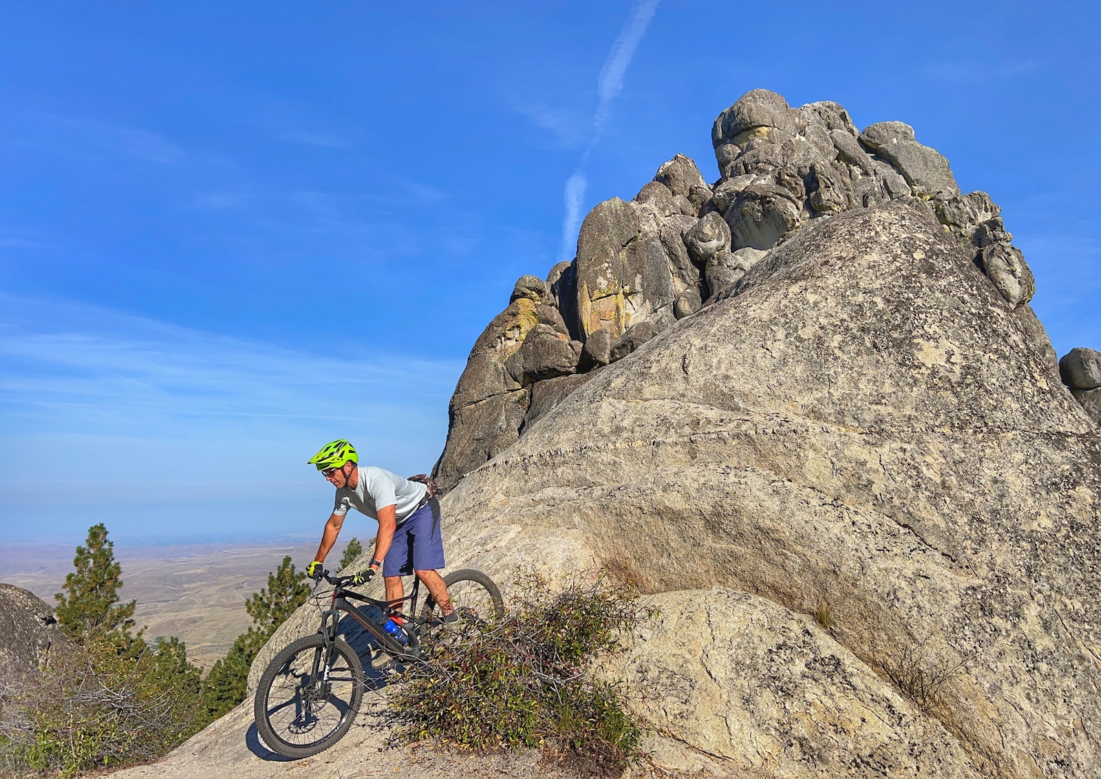 Mountain biker riding on rocky terrain beside large granite rock formation with valley views.