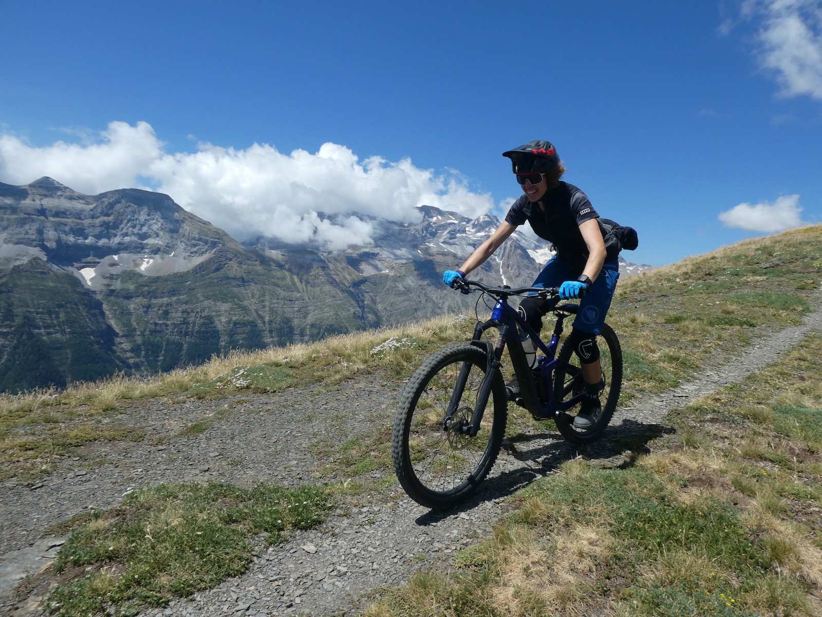 A rider on a gravel trail in alpine terrain with snow-capped peaks in the background.