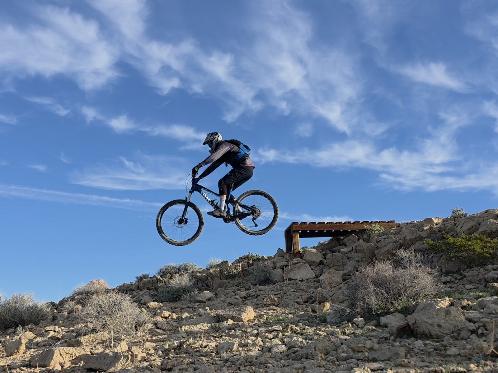 A mountain biker airborne off a wooden jump on rocky desert terrain.