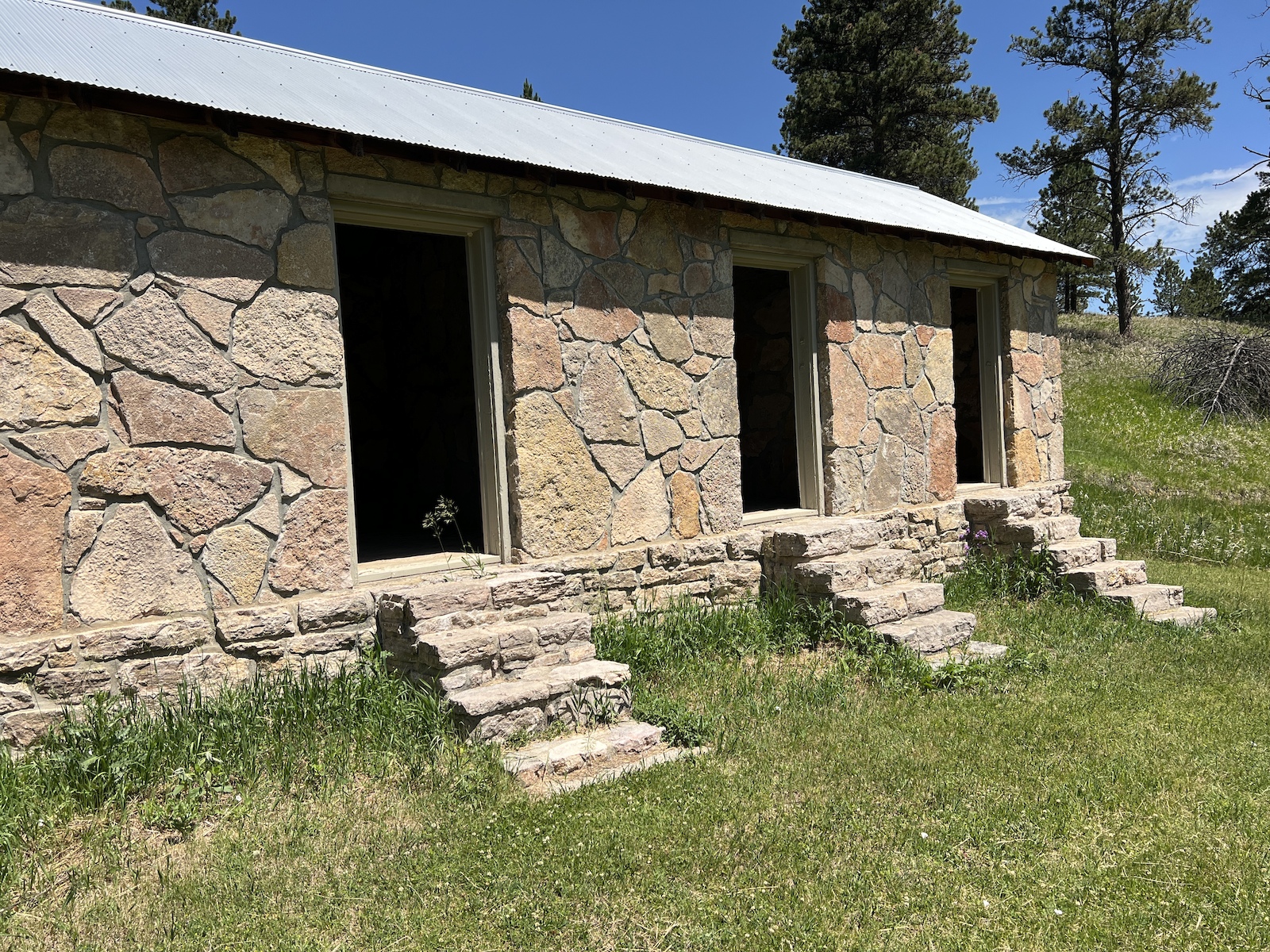 A historic stone building with multiple doorways and a metal roof surrounded by grass and trees.