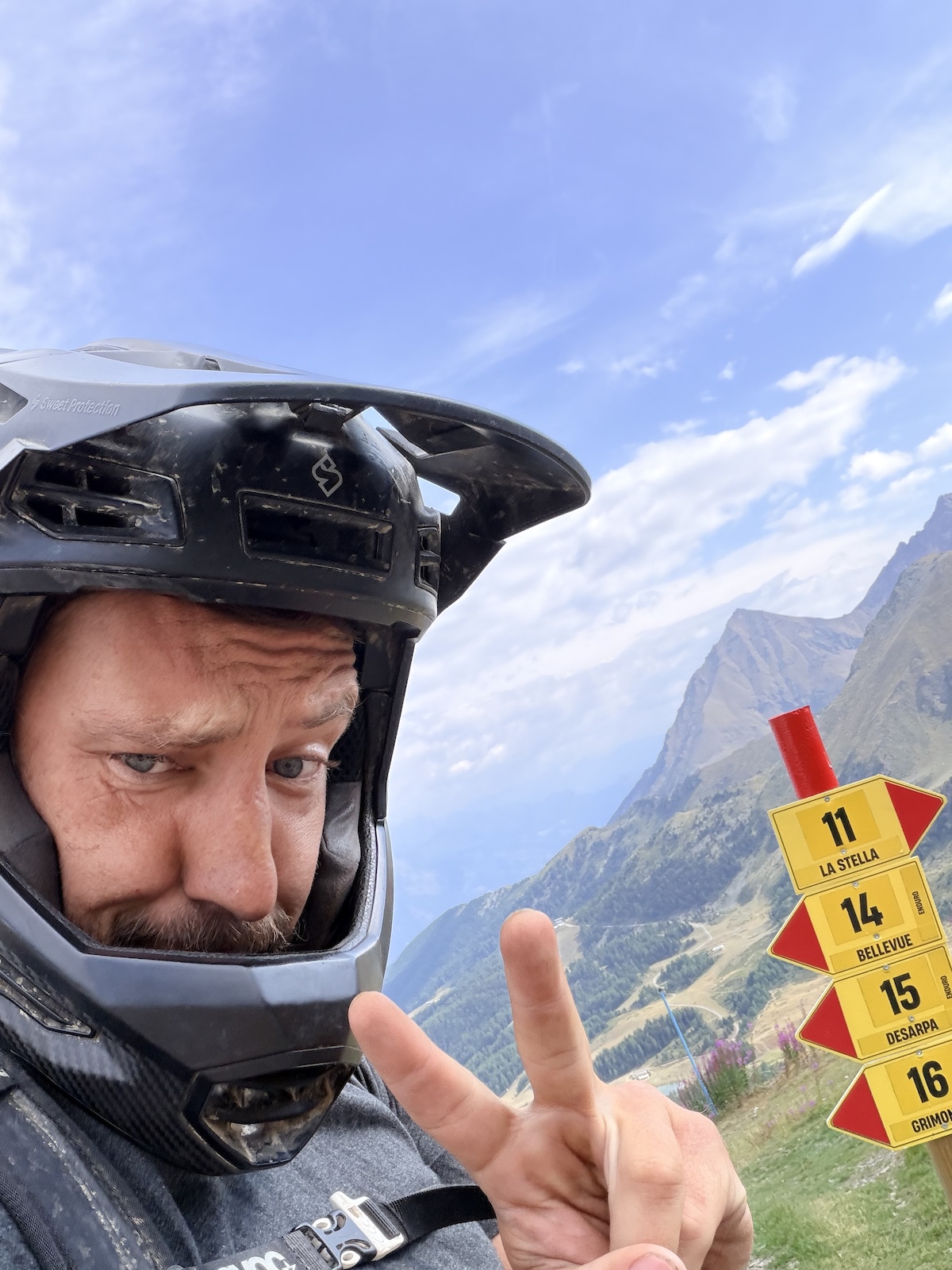 A person wearing a black helmet making a peace sign with mountains and trail markers visible.
