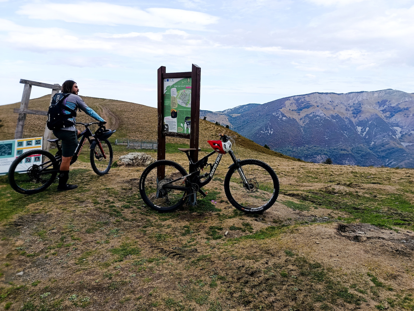 Two cyclists standing at trailhead with map sign and mountain views.